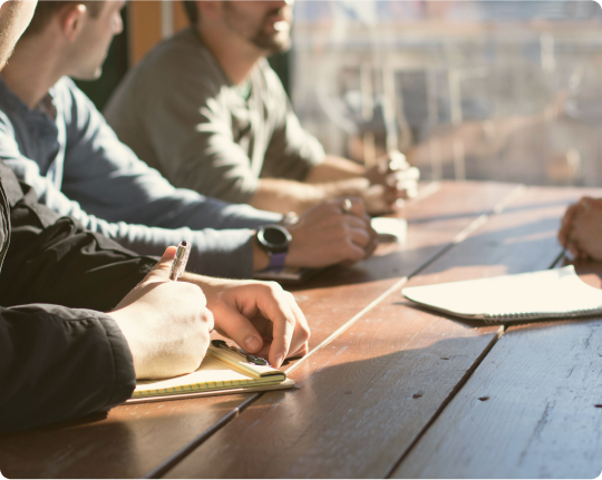 Group of people seated at a wooden table, engaged in a discussion and taking notes.